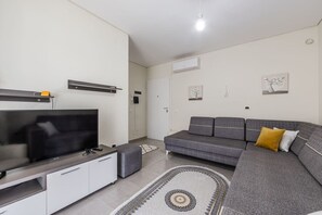 Een moderne woonkamer met een flatscreen televisie, een grijze bank, witte kasten en een vloerkleed met een patroon.
