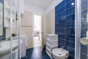 Moderni kylpyhuone, jossa on pesuallas, wc ja bidee, lasinen suihkuseinä ja näkymä olohuoneeseen.