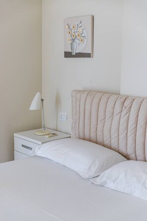 Een net opgemaakt bed met witte lakens, een bedlampje, een klein nachtkastje en een aan de muur hangend bloemenstillife.
