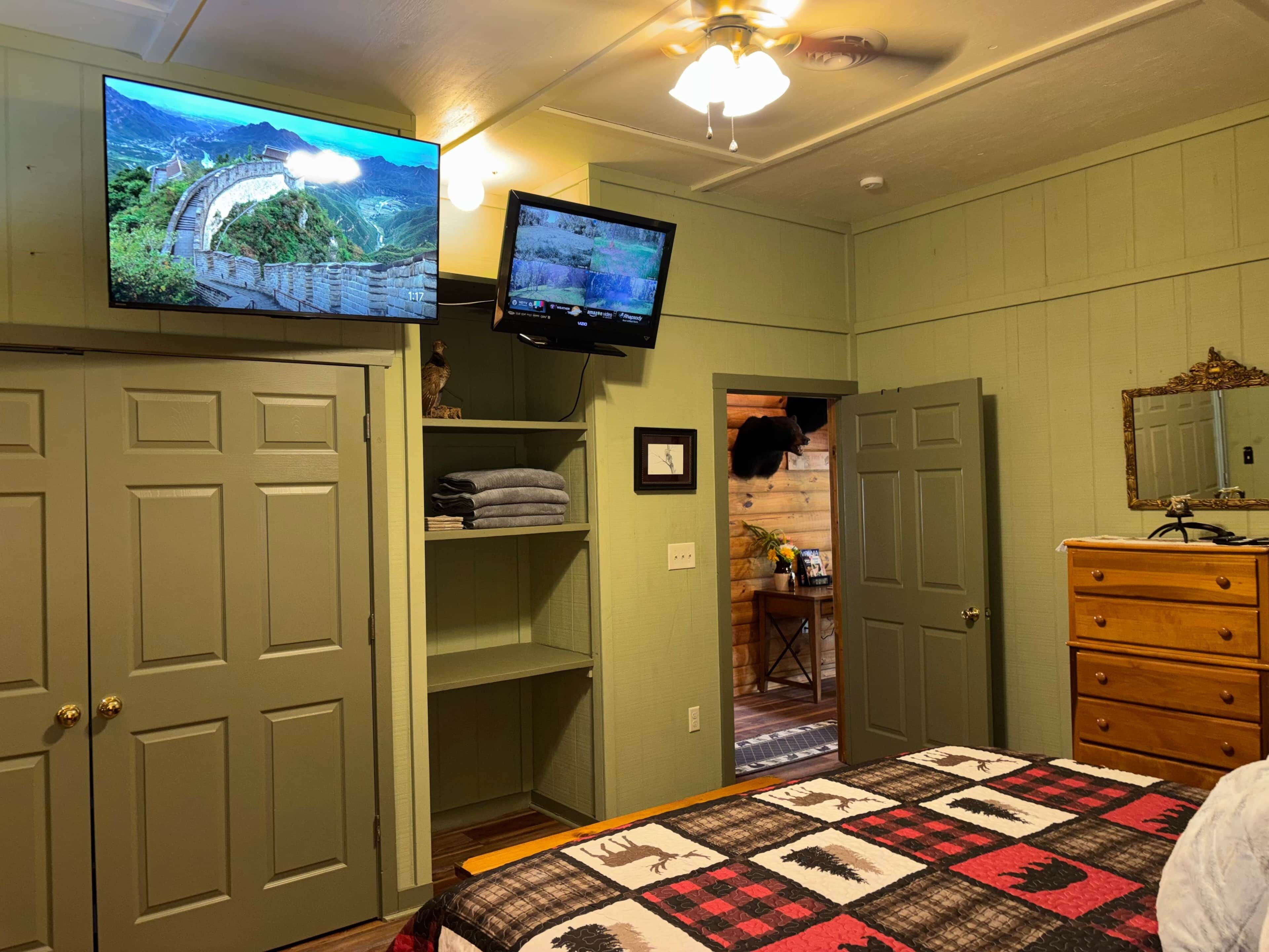 Quarto com duas televisões fixadas na parede, uma cama com colcha, cômoda e porta que dá para outro ambiente.