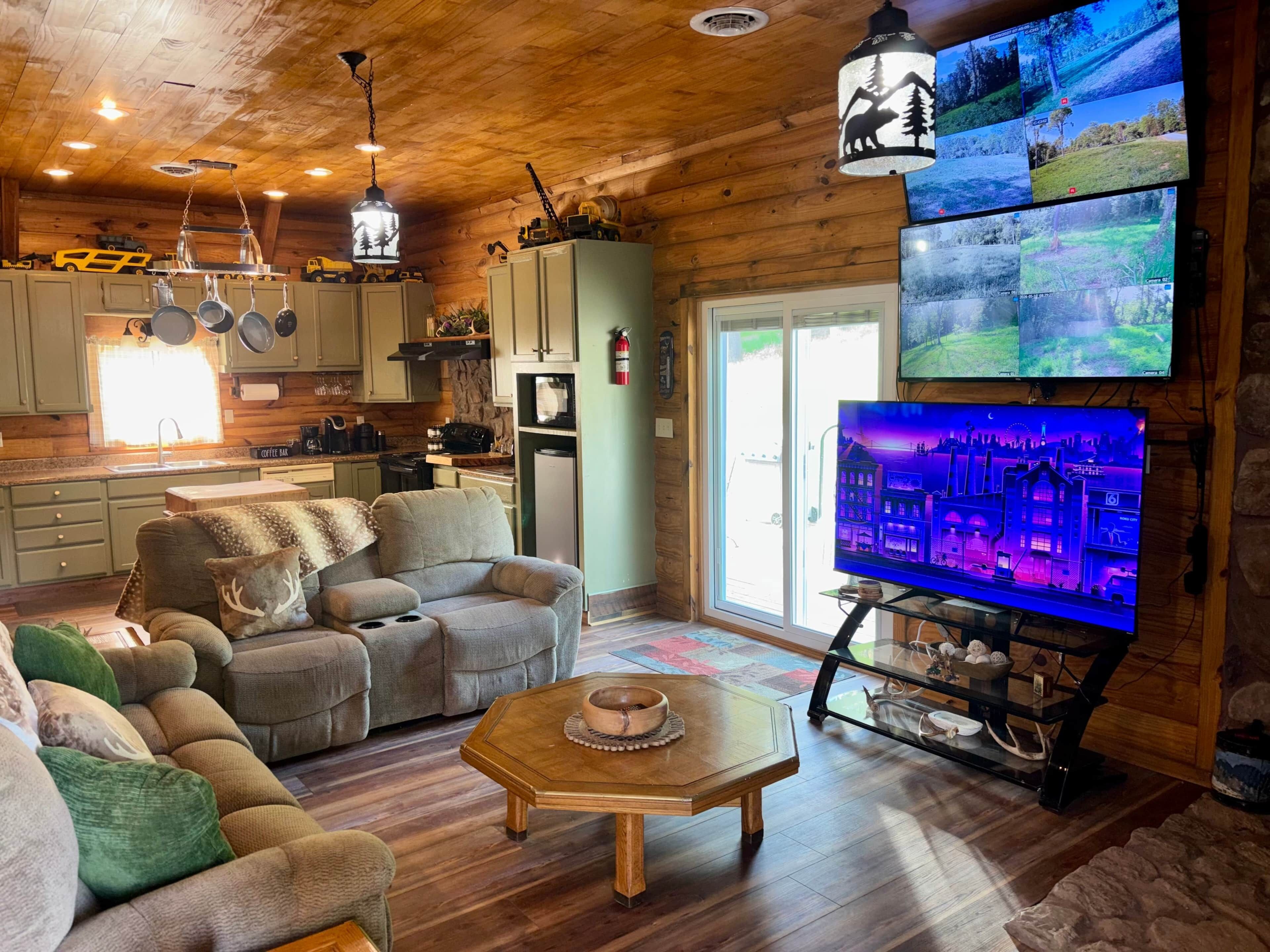 Een gezellig chaletinterieur met een zithoek, keuken en een groot televisiescherm waarop een stadsgezicht te zien is.
