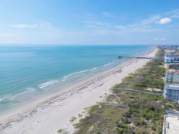 Eine Luftaufnahme eines Strandes mit einem Steg, der sich ins Meer erstreckt, neben einer Küstenstraße und Gebäuden.