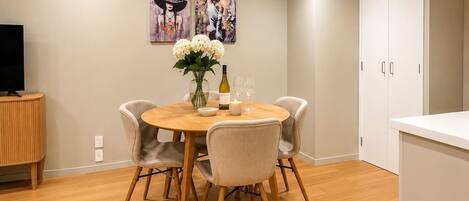 Uma sala de jantar com uma mesa redonda, quatro cadeiras, um vaso com flores, uma garrafa e copos de vinho.
