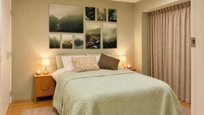 Ein Schlafzimmer mit einem Bett, zwei Nachttischlampen, einem Nachttisch, einer Wand mit mehreren gerahmten Bildern und einem Fenster mit Vorhängen.