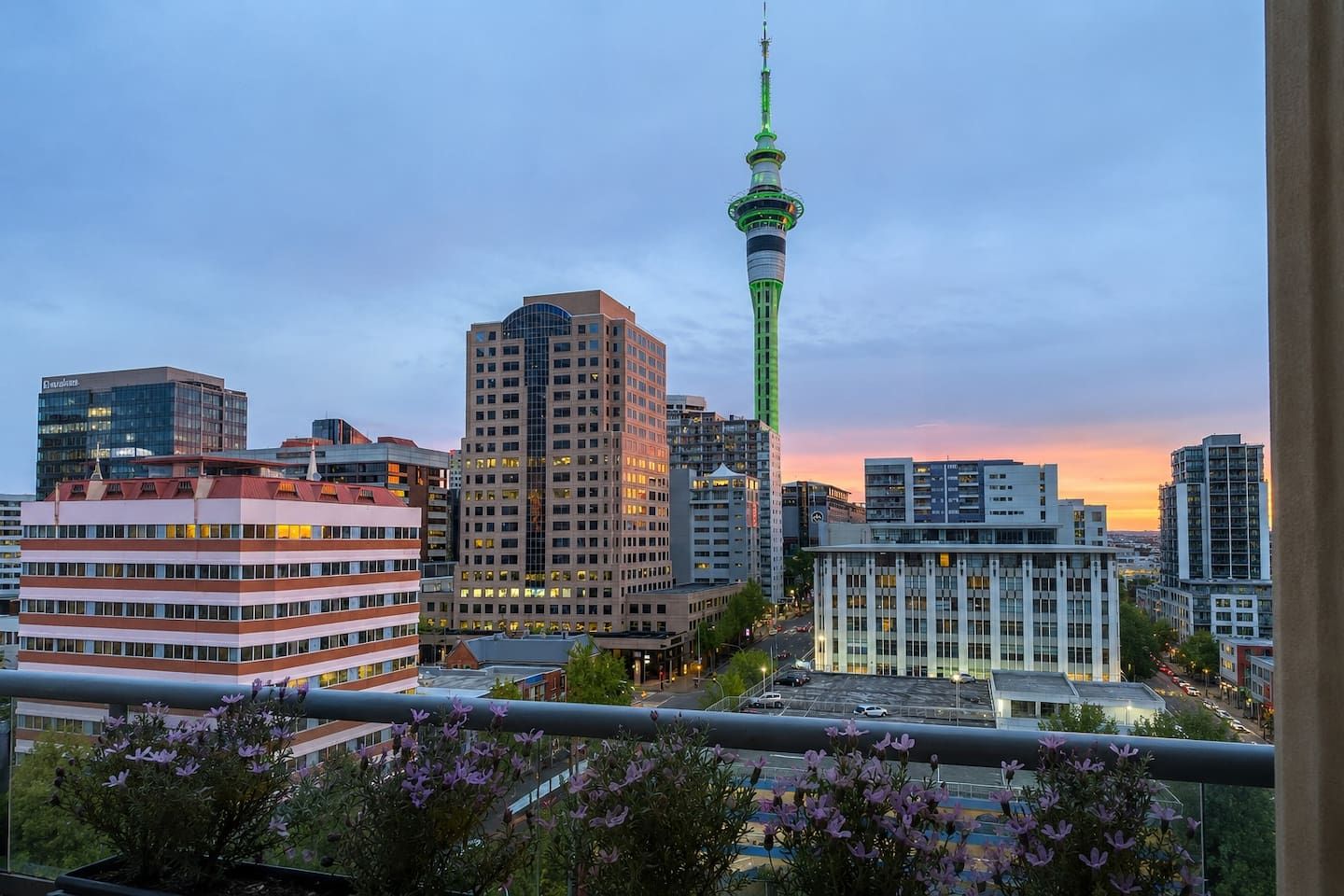 A cityscape with modern buildings, a prominent tower, and a balcony with flowers.
