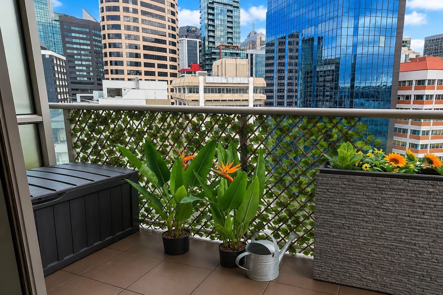 A balcony with potted plants, a metal watering can, and a view of a cityscape with modern buildings.