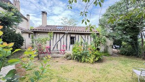 Ein rustikales Haus mit Schornstein, ein kleiner Garten mit Tisch und Stühlen sowie eine Metallbank.