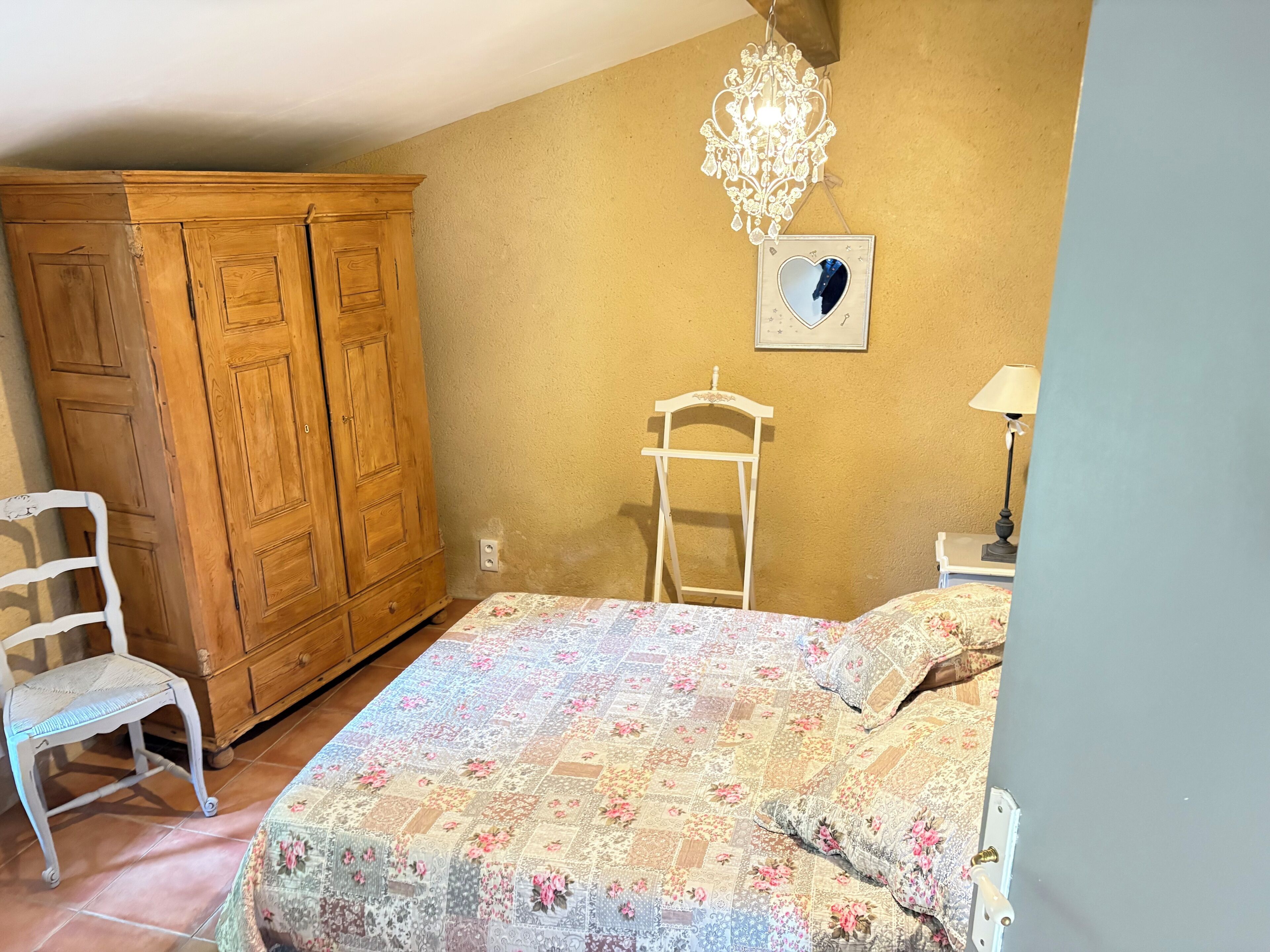 A bedroom with a wooden wardrobe, a white chair, a bed with a floral quilt, a chandelier, and a wall-mounted picture.