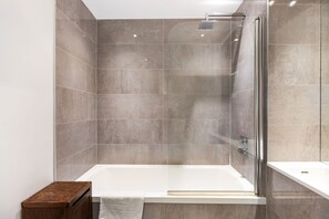 A modern bathroom with a glass-enclosed shower, a white bathtub, and a wooden bench. - GuestReady - Chic Urban Retreat with Balcony Views (London)