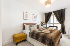 A bedroom with a bed, a yellow ottoman, a window with curtains, and two framed pictures on the wall. - GuestReady - Chic Urban Retreat with Balcony | 20min Oxford Circus (London)