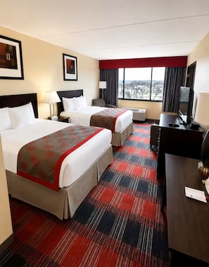 A hotel room with two beds, a desk with a TV, and a large window.