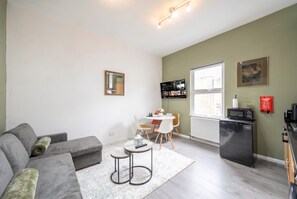 Ein kompakter Wohnraum mit grauem Sofa, einem kleinen Esstisch mit Stühlen, einer Küchenzeile mit Mikrowelle und einem Fenster mit Jalousien.