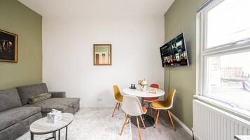 A living room with a sofa, a round table with chairs, a TV mounted on the wall, and a window with a view.