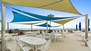 An outdoor patio with white metal tables and chairs, shaded by blue and beige canopies, overlooking the sea. - Gulf View Condo at Dunes of Panama with Pool (Panama City Beach)