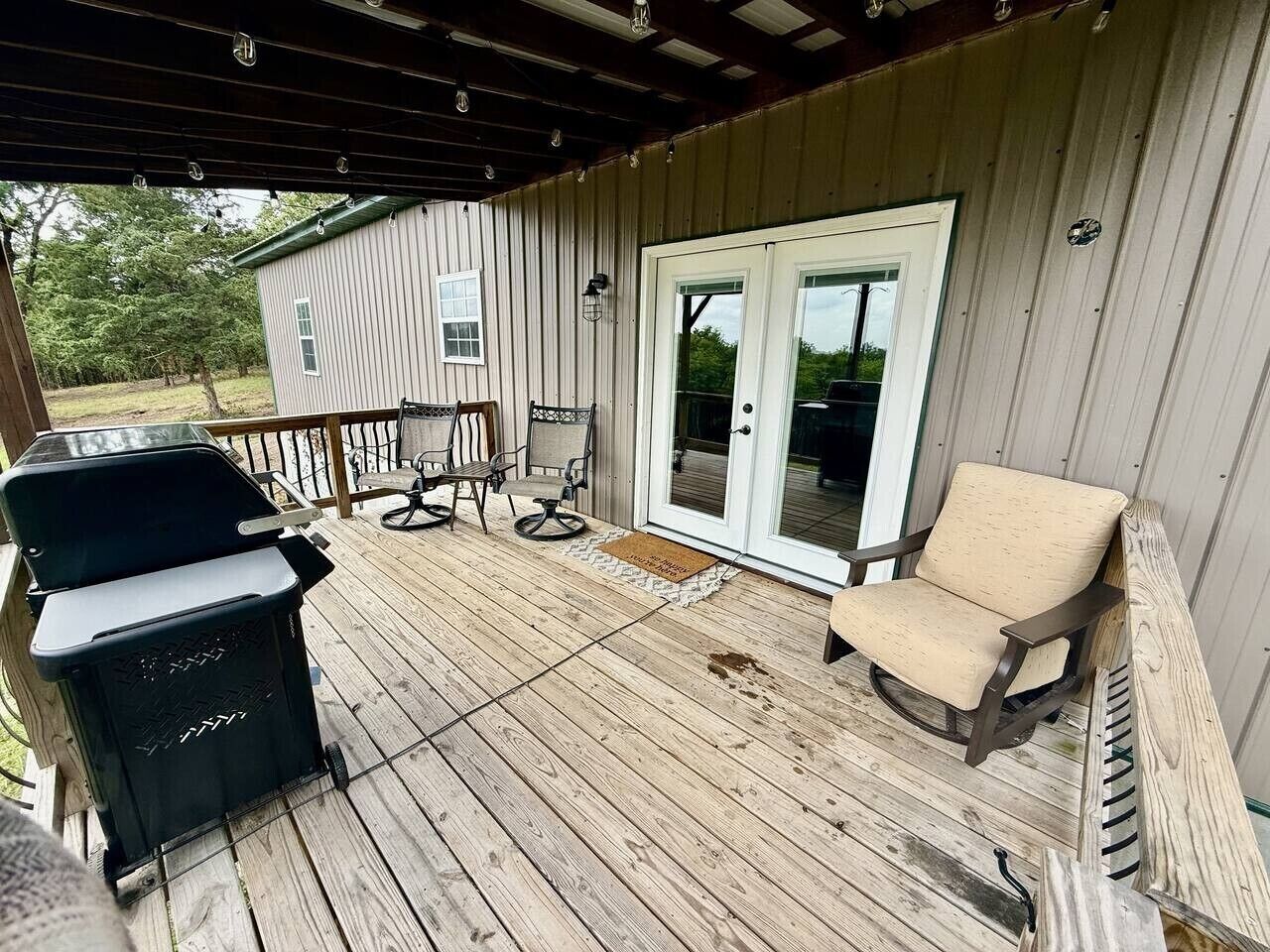 Een overdekt terras met een barbecue, twee stoelen en een schuifdeur naar een gebouw.