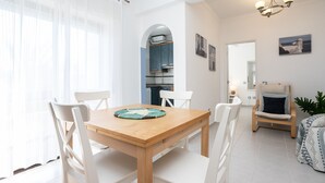 Uma sala de jantar com mesa de madeira e cadeiras brancas, uma cozinha com armários azuis e uma sala de estar com uma cadeira e uma planta.