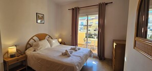 Una habitación de hotel con cama, mesita de noche con lámpara, un cuadro en la pared y vistas al balcón exterior.