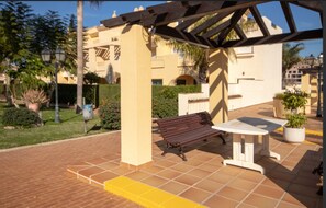 An outdoor patio with a bench and table, surrounded by potted plants and a building with balconies.
