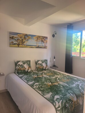 Ein Schlafzimmer mit einem Bett, einem Palmen-Gemälde, einem Nachttisch, einer Lampe und einem Fenster mit Vorhängen.