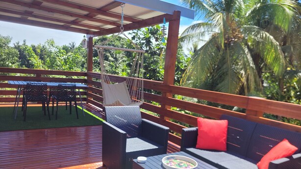 An outdoor deck with a wooden floor, a hammock, wicker furniture, and a table with a bowl. - Pom Kanel Vacation Rental with Unobstructed Views (Sainte-Anne)