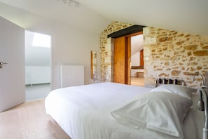 Une chambre avec un mur en pierre, un lit avec une literie blanche, un radiateur et une porte donnant sur une autre pièce.