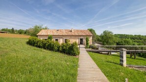 Un bâtiment rustique avec un toit en tuiles, un chemin en bois menant à l’entrée et un champ verdoyant en arrière-plan.