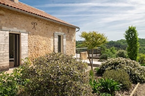 Un bâtiment en pierre doté de deux portes, entouré d’arbustes bien entretenus et d’un arbre.