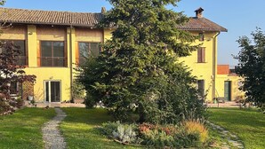 A yellow building with a tiled roof, a gravel path, and a large evergreen tree. - Il Casale: A Charming Country House in the Monferrato Hills (Colcavagno)