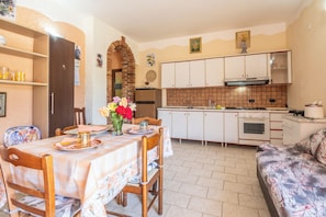 A dining area with a table set for two, a sofa, and a kitchen with white cabinets and a tiled backsplash. - Villa Baronia (Stampella)
