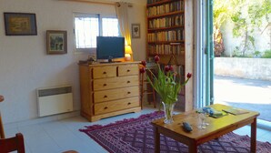 Et værelse med en trækommode, et fjernsyn, en reol, et vindue med gardiner, en vase med blomster og et bord med en fjernbetjening og glas.