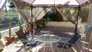 En overdækket terrasse med et bord og stole, omgivet af krukker med planter og et hegn.