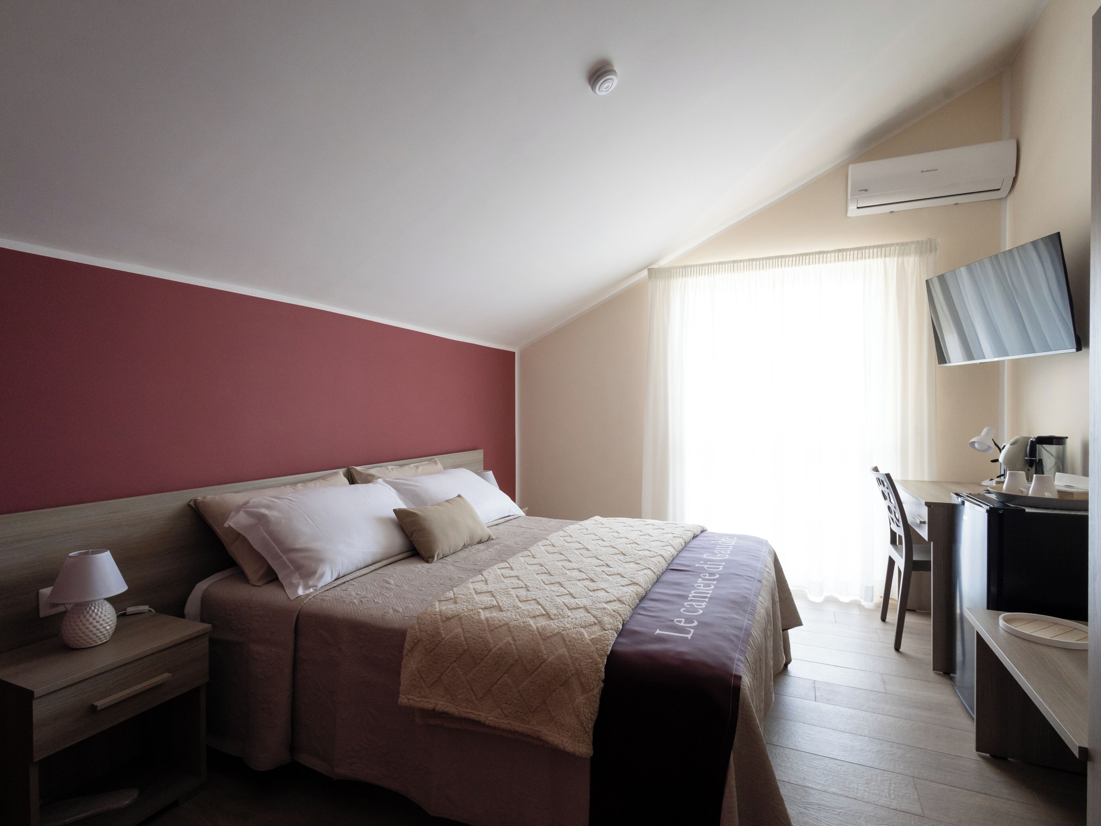 Una habitación de hotel con cama, mesita de noche, lámpara, escritorio con silla y equipo de aire acondicionado.