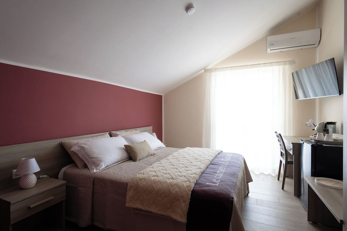 Ein Hotelzimmer mit Bett, Nachttisch, Lampe, Schreibtisch mit Stuhl und einer Klimaanlage.
