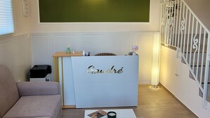 Una sala de estar con un sofá color beige, una mesa de centro blanca, una recepción con letrero 'Gauthé', una pared de acento verde y una escalera con barandales blancos.