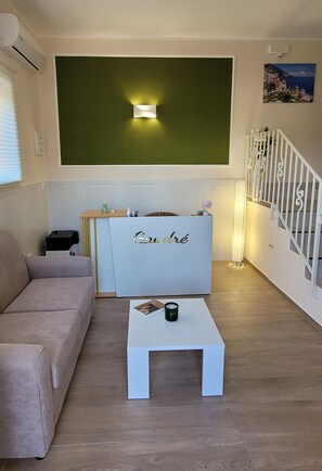 Sala de estar com sofá bege, mesa de centro branca, balcão de recepção com a identificação 'Gauthé', parede de destaque verde e escada com corrimão branco.