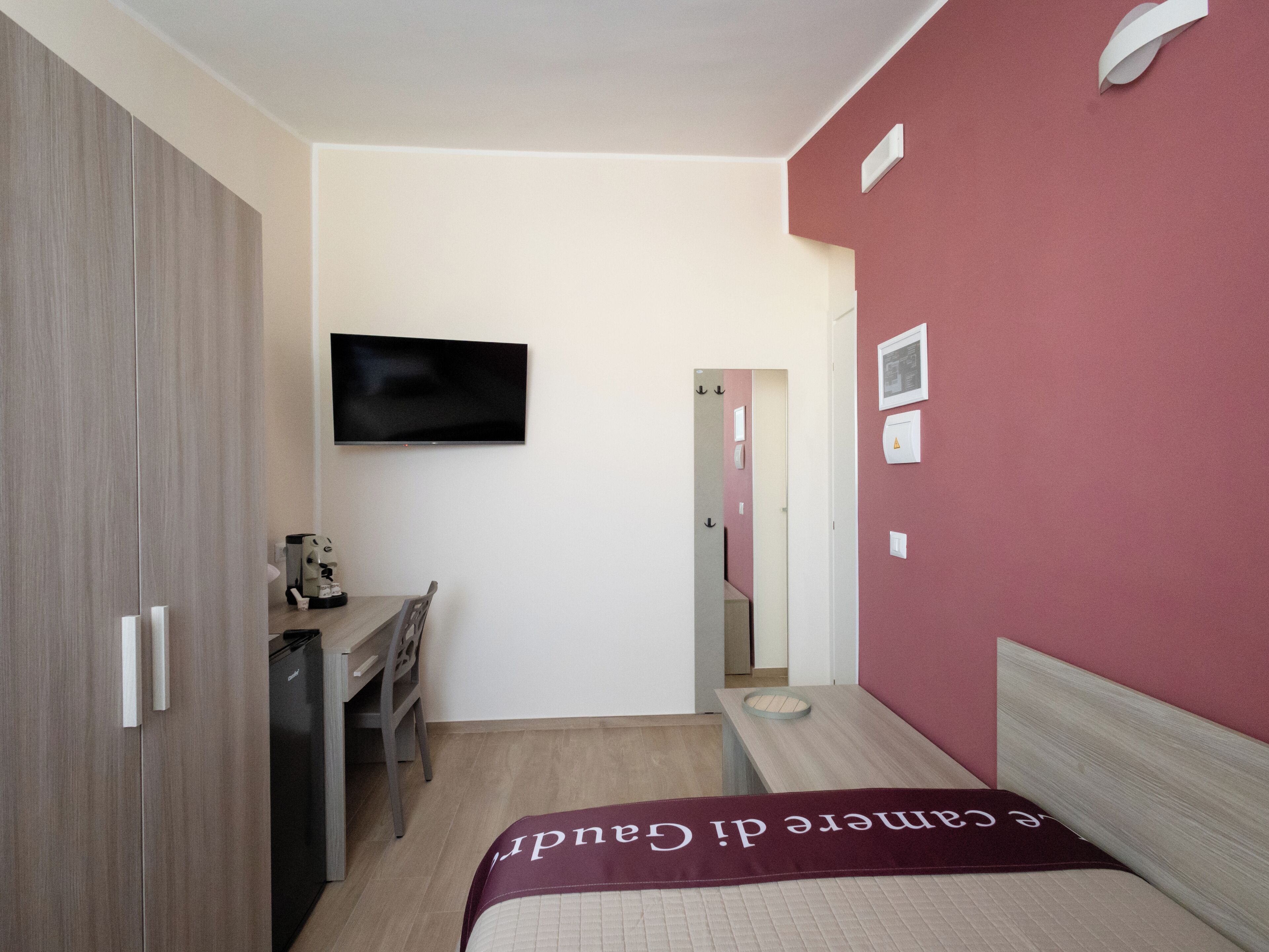 A compact hotel room with a bed, a desk, a chair, a television mounted on the wall, and a door leading to another room.