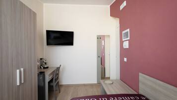 A compact hotel room with a bed, a desk, a chair, a television mounted on the wall, and a door leading to another room.