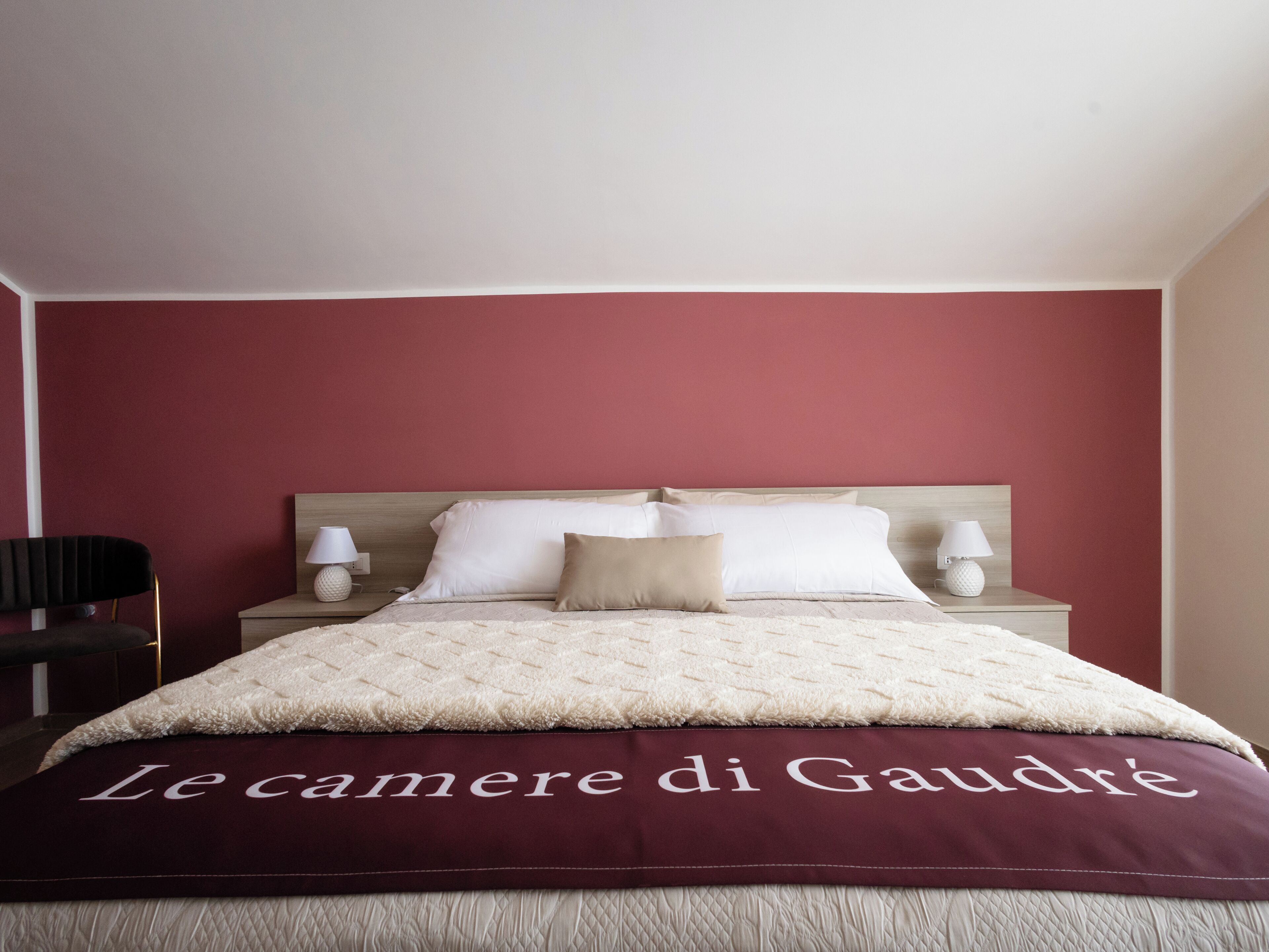 A neatly made bed with a red headboard, white pillows, and a beige blanket. There are two bedside lamps on either side of the bed.