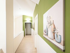 Um corredor com parede verde, assoalho de madeira e um quadro de vasos à direita.