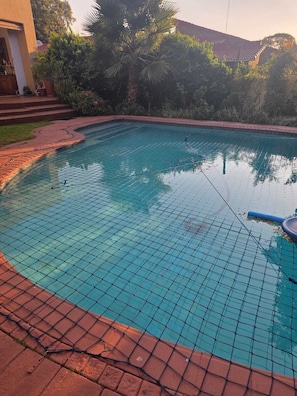 A swimming pool with a tiled edge, surrounded by greenery and a building. - Sunny sanctuary in the heart of Gallo Manor (Sandton)