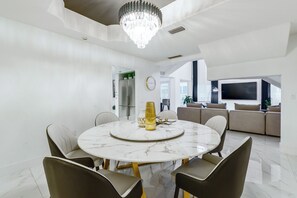 A modern dining area with a marble table, six chairs, a chandelier, a refrigerator, and a TV in the background.