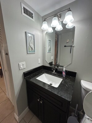 A bathroom with a black countertop sink, a mirror with lights, a framed giraffe picture, and a vent on the wall. - The Native-Vintage Florida Vibe in the Oldest City (St. Augustine)
