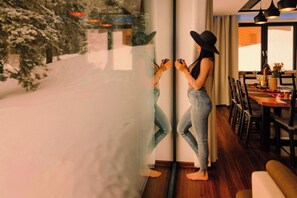 Eine Person steht an einem Fenster mit Blick auf Schnee, ein Essbereich mit Stühlen und einem Tisch, und die Person spiegelt sich im Glas.