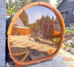 Ein runder Spiegel, der einen Garten mit einer Holzhütte, einem Blumentopf und einem Metallstuhl reflektiert.