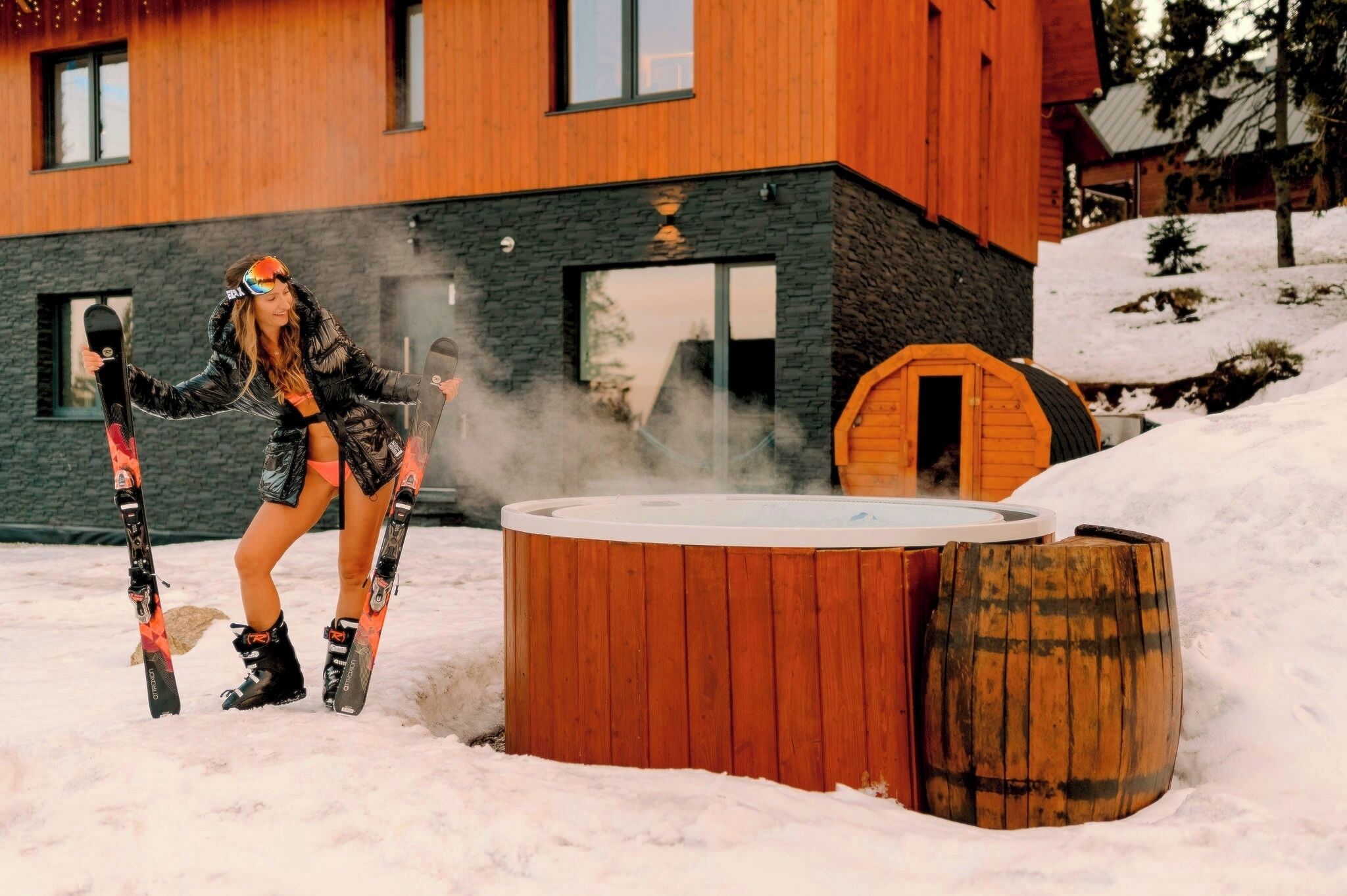 A wooden building with a stone facade, a hot tub, and a wooden barrel in the snow.
