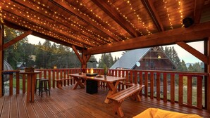 A wooden deck with a fire pit, tables, and benches, overlooking a forested area. - Mountain Chalet for 20 with Wellness, Sauna & Hot Tub — Ski-in/Ski-out (Martin)