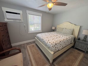 Quarto com cama, ventoinha no teto, ar condicionado, janela com persianas, mesa de cabeceira com abajur e cômoda.