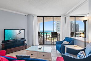 Ein modernes Hotelzimmer mit Fernseher, einem Glastisch, einer Couch und einem Balkon mit Blick auf das Meer und Gebäude.