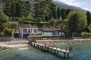 Ein Haus am Wasser mit einer Holzsteganlage, einem zweistöckigen Gebäude mit Balkon und einer Bergkulisse im Hintergrund.