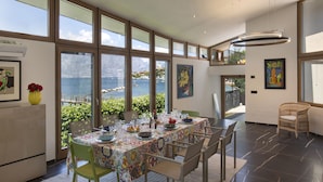 A dining area with a table set for four, large windows with a view of the sea, and a high ceiling with recessed lighting. - Villa Locchi with private Pier (Malcesine)